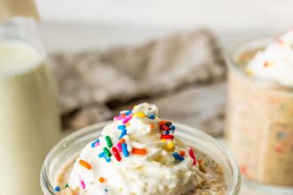 A glass jar filled with birthday cake overnight oats, topped with whipped cream and colorful sprinkles, with a bottle of milk and another jar of oats in the background.