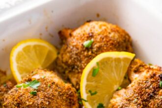 Six baked Greek chicken thighs in a white baking dish. The chicken is topped with fresh parsley and lemon slices.