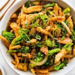 Chicken and broccoli stir fry in a white bowl, garnished with sesame seeds and green onions, with wooden chopsticks beside it.